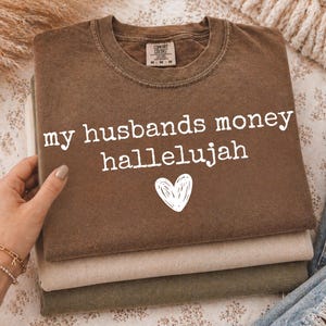 May include: A brown crewneck t-shirt with the white text "my husbands money hallelujah" and a white heart graphic printed on the front. The shirt features a Comfort Colors brand label at the inner neckline and is stacked atop other folded shirts.