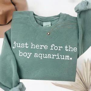 以下が含まれることがあります： 「just here for the boy aquarium」の白文字が入ったセージグリーンのスウェットシャツ。クルーネックと、ロールカフスの長袖。小さなペンダントが付いたゴールドのネックレスが見えます。
