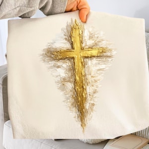 Könnte beinhalten: Cremefarbene Decke mit einem großen, strukturierten goldenen Kreuzdesign. Das Kreuz ist der Mittelpunkt, mit pinselstrichartigen Details. Die Decke wird von einer Person gehalten, die das Design präsentiert.