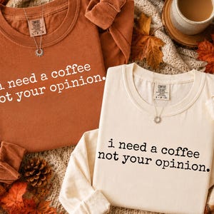 May include: Two long-sleeve crewneck shirts, one in burnt orange with white text and one in cream with black text. Both shirts feature the phrase "i need a coffee not your opinion." and a Comfort Colors brand label at the inner neckline.