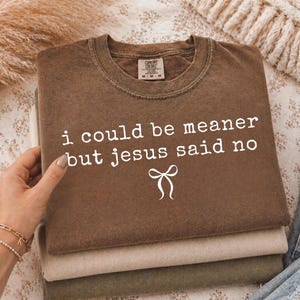 Op de afbeelding: Een stapel van drie Comfort Colors t-shirts in bruin, beige en olijfgroen. Het bovenste shirt heeft de witte tekst "i could be meaner but jesus said no" en een strik-afbeelding. De shirts zijn opgevouwen.