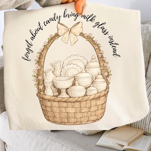 May include: A beige tote bag featuring a detailed illustration of a woven basket filled with vintage milk glass. The basket is adorned with a floral garland and a bow. The text reads "forget about candy bring milk glass instead."
