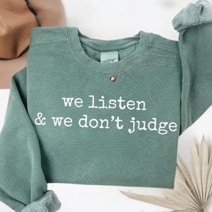 Puede incluir: Sudadera verde salvia con el texto blanco "we listen & we don't judge". Un collar dorado con un pequeño colgante está drapeado en la parte delantera. Las mangas están dobladas. Un sombrero de tonos neutros es visible en el fondo.