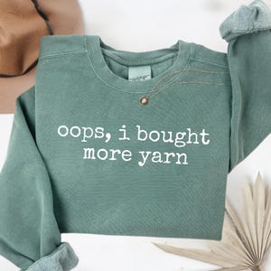 Puede incluir: Una sudadera verde salvia con la frase "oops, i bought more yarn" en letras blancas. Un collar dorado con un colgante descansa sobre la camisa. Las mangas están dobladas y se ve parte de un sombrero marrón en el fondo.