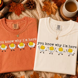 May include: Two crew neck t-shirts, one in burnt orange and one in cream, each featuring the text you know why i'm here and a graphic of four cartoon deviled eggs with smiling faces and stick arms and legs printed on the front chest area.