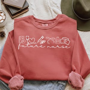 May include: A terracotta-coloured sweatshirt featuring white text and medical-themed graphics. The text reads "future nurse" with illustrations of a stethoscope, heart, syringe, bandage, and other medical symbols. The sleeves are cuffed.