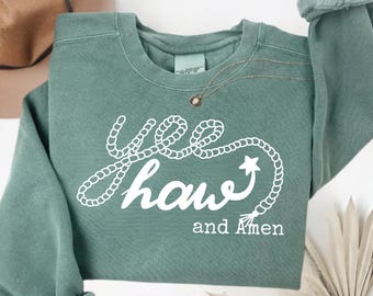 Puede incluir: Una sudadera verde salvia con las palabras "Yee Haw" en blanco, con un diseño de cuerda, y "and Amen" debajo. Se ve un collar dorado. Las mangas están dobladas. Un sombrero de vaquero es parcialmente visible en el fondo.