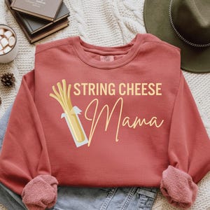 以下が含まれることがあります： テラコッタ色のスウェットシャツ。ライトベージュの筆記体で「STRING CHEESE Mama」と書かれています。ストリングチーズのグラフィックも特徴です。スウェットシャツは長袖で、クルーネックです。