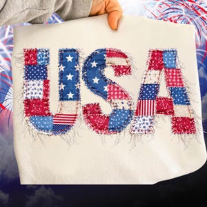 Puede incluir: Una tela color crema con la palabra "USA" aplicada en patchwork rojo, blanco y azul. Las letras están hechas de varios patrones patrióticos, incluyendo estrellas, rayas y paisley. Los bordes tienen un detalle deshilachado y cosido.