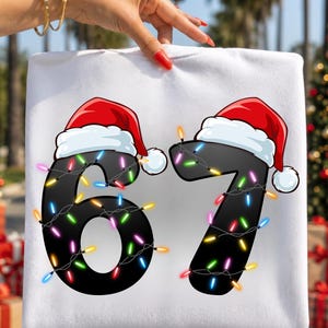May include: A white sweatshirt featuring a graphic of the number 67 in black. Each digit is topped with a red and white Santa hat and wrapped in a string of colourful Christmas lights in shades of blue, yellow, green, pink, and orange.