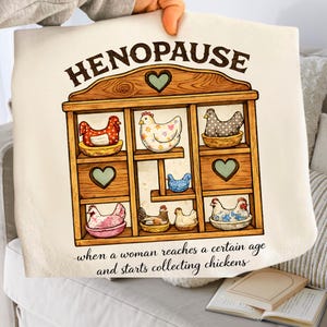 May include: Cream-colored decorative pillow with a wooden shelf design featuring cartoon chickens in various patterns and colors. The text "HENOPAUSE" is at the top, with the phrase "when a woman reaches a certain age and starts collecting chickens" at the bottom.