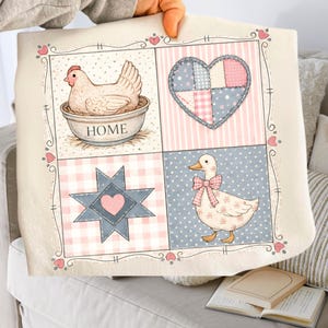 Puede incluir: Una manta crema con diseño de patchwork. Presenta una gallina, un corazón, una estrella y un pato con un lazo rosa. La manta tiene un borde decorativo con corazones. La palabra "HOME" es visible.