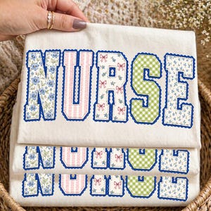 May include: A cream-colored t-shirt featuring the word NURSE in large block letters. Each letter has a distinct pattern: blue floral, pink stripes, white with pink bows, green gingham, and blue floral, all outlined in a dark blue scalloped stitch.