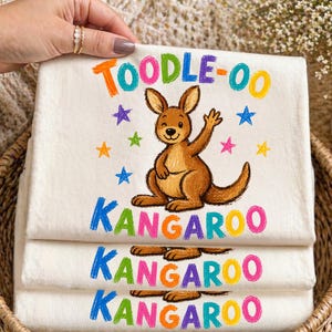 May include: Off-white cotton tea towels with a colorful kangaroo illustration. The towels feature the words "TOODLE-OO KANGAROO" in rainbow colors, along with star accents. The kangaroo is brown and winking.