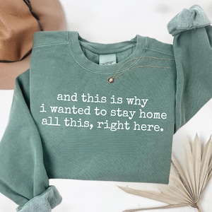 May include: A sage green sweatshirt with the text "and this is why I wanted to stay home all this, right here." A gold necklace with a small pendant is draped on the front. The sleeves are cuffed.
