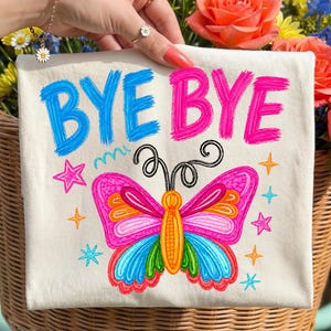 Può includere: Borsa tote color crema con le parole "BYE BYE" in blu e rosa, e un'illustrazione di farfalla colorata. La farfalla ha ali rosa, arancioni, blu e verdi. La borsa è decorata con stelle e linee ondulate.