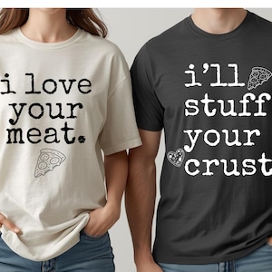 May include: Two t-shirts, one cream and one dark gray. The cream shirt says "i love your meat" with a pizza slice graphic. The dark gray shirt says "i'll stuff your crust" with a pizza slice and heart graphic. Both shirts are short-sleeved.