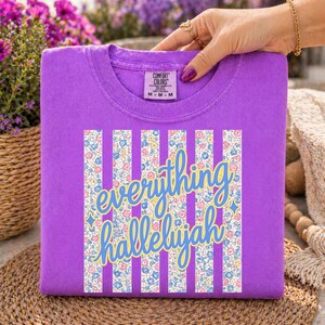 May include: A purple Comfort Colors t-shirt with the words "everything hallelujah" in a blue and yellow script font. The design includes vertical white stripes with a floral pattern. The shirt is a medium size.