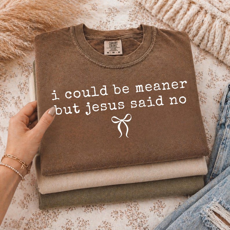 Op de afbeelding: Een stapel van drie Comfort Colors t-shirts in bruin, beige en olijfgroen. Het bovenste shirt heeft de witte tekst "i could be meaner but jesus said no" en een strik-afbeelding. De shirts zijn opgevouwen.