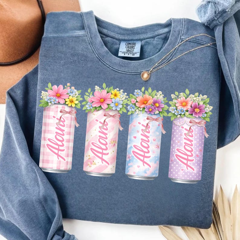 Pode incluir: Um moletom azul com um design floral. O design inclui quatro latas decorativas com o nome "Alani" impresso. Cada lata &eacute; adornada com um padr&atilde;o diferente, flores e uma fita.