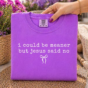 Op de afbeelding: Een paars Comfort Colors T-shirt met de witte tekst "i could be meaner but jesus said no" en een strik. Het T-shirt heeft een ronde hals en is opgevouwen. Het shirt wordt door een hand vastgehouden.