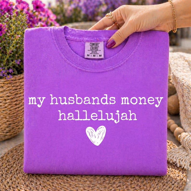 May include: A purple short-sleeved t-shirt featuring the white text 'my husbands money hallelujah' printed above a white hand-drawn heart graphic. The shirt has a Comfort Colors brand label on the inside collar indicating a size medium.