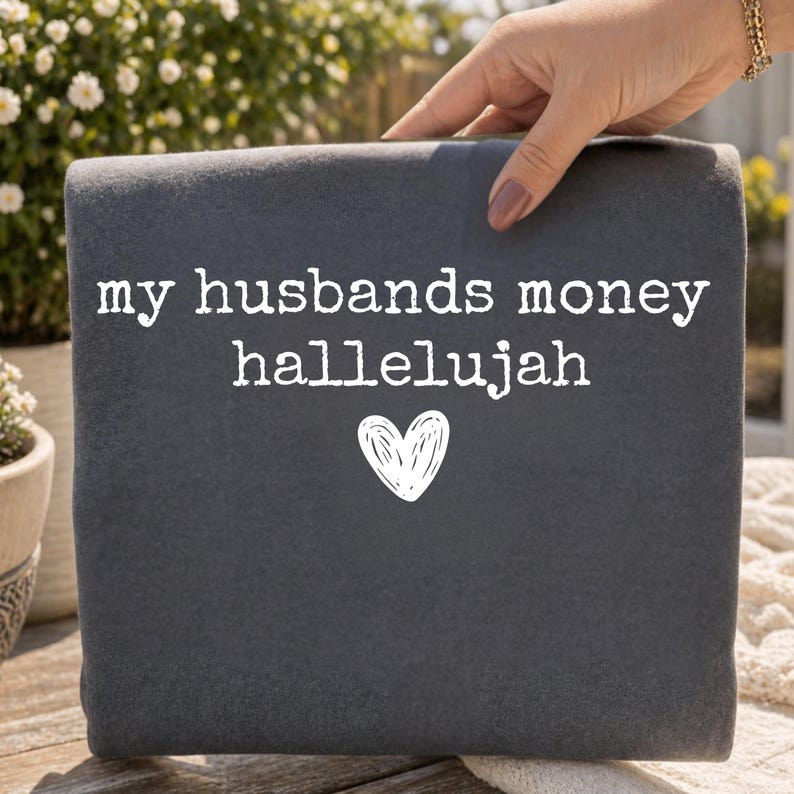 May include: A dark gray folded fabric item featuring the white printed text "my husbands money hallelujah" arranged in three lines, with a simple white hand-drawn heart icon centered below the text on the front surface of the material.