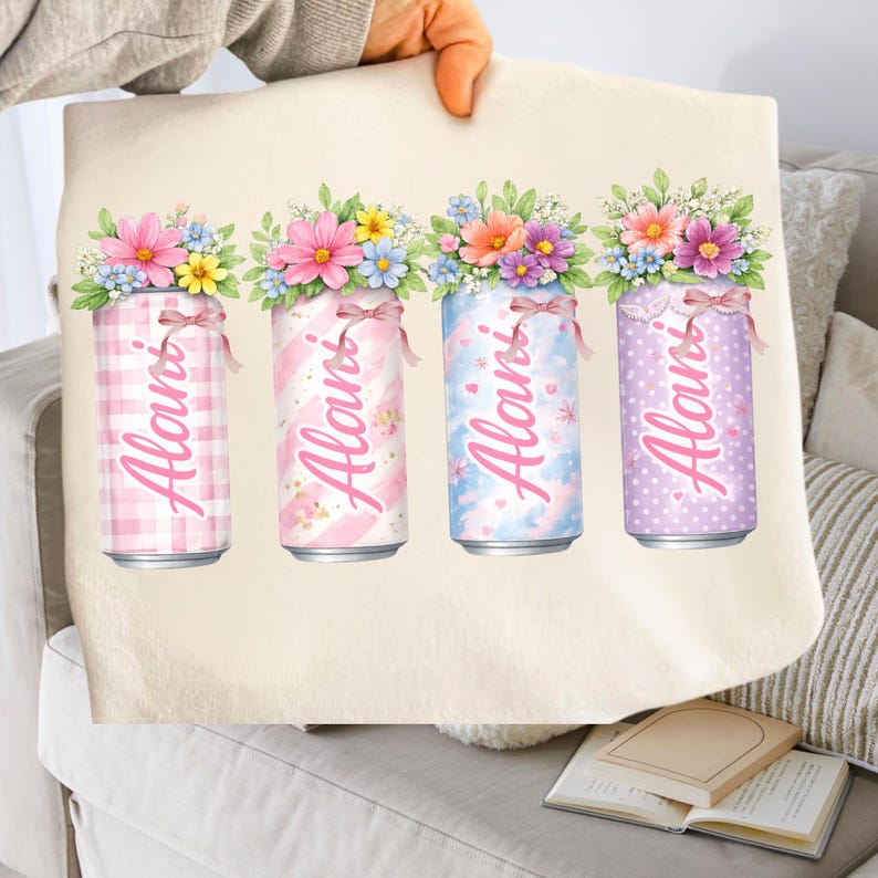 Pode incluir: Uma sacola bege com quatro latas decorativas com arranjos florais. Cada lata tem uma cor pastel e um padr&atilde;o diferente, com o nome "Alani" em script rosa. As latas s&atilde;o cobertas com flores rosa, amarelas e azuis.