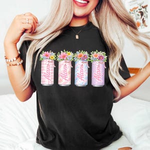 Pode incluir: Camiseta preta com quatro latas decorativas com arranjos florais. Cada lata tem um padr&atilde;o diferente e o nome "Alani" impresso. As latas s&atilde;o rosa, azul e roxo.