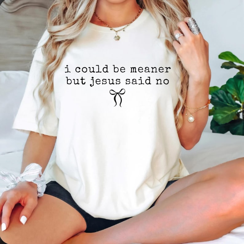 Op de afbeelding: Cr&egrave;mekleurig T-shirt met de zwarte tekst "i could be meaner but jesus said no" en een zwarte strik-afbeelding. Het shirt is een casual stijl met korte mouwen.