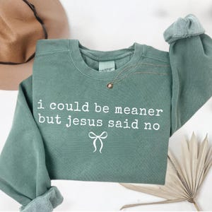 Op de afbeelding: Saliegroen sweatshirt met de witte tekst "i could be meaner but jesus said no". Een gouden ketting met een kleine hanger is over de voorkant gedrapeerd. De mouwen zijn opgerold. Een hoed is zichtbaar op de achtergrond.