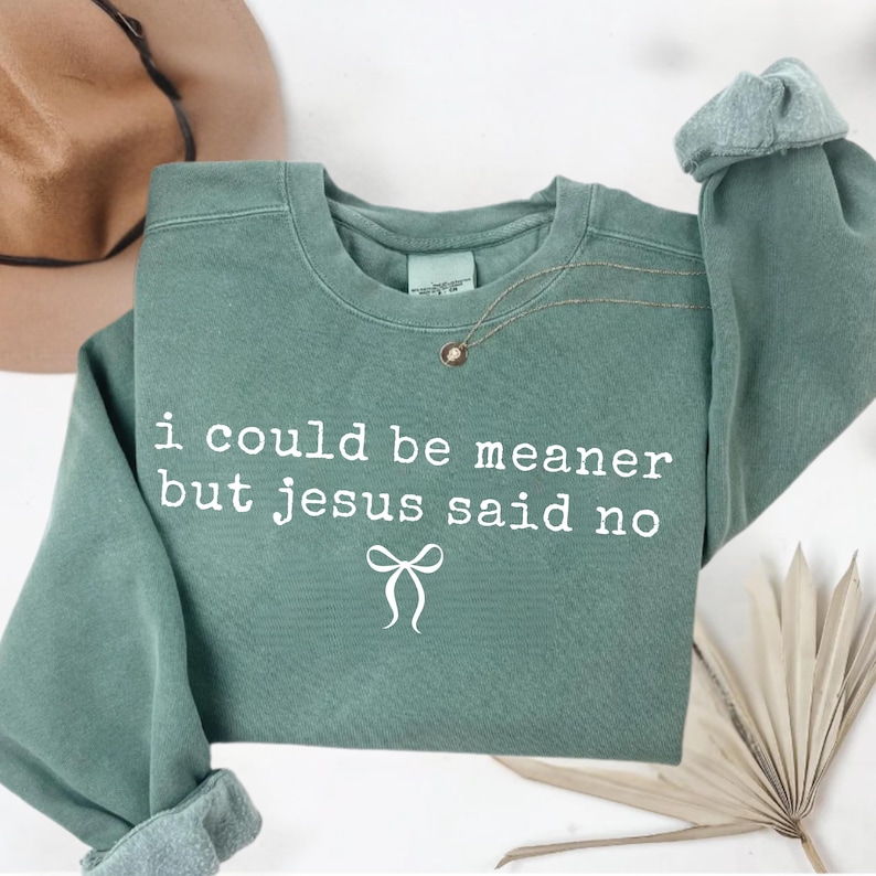 Op de afbeelding: Saliegroen sweatshirt met de witte tekst "i could be meaner but jesus said no". Een gouden ketting met een kleine hanger is over de voorkant gedrapeerd. De mouwen zijn opgerold. Een hoed is zichtbaar op de achtergrond.