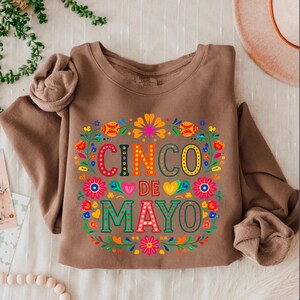 May include: A brown sweatshirt featuring the text "CINCO DE MAYO" in vibrant colours, surrounded by a floral wreath. The design incorporates flowers in red, orange, yellow, and blue, celebrating the Mexican holiday.