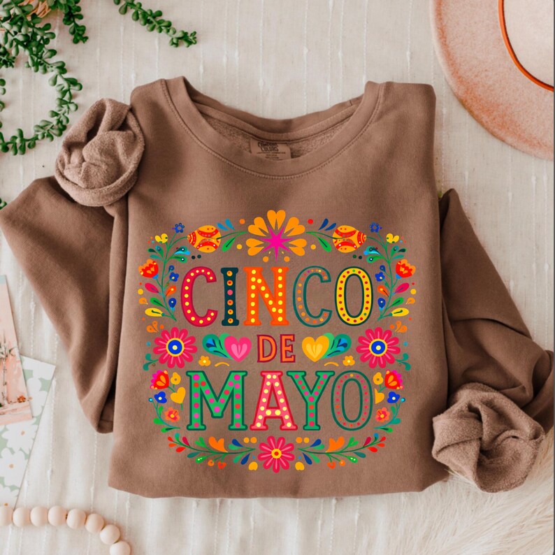 May include: A brown sweatshirt featuring the text "CINCO DE MAYO" in vibrant colours, surrounded by a floral wreath. The design incorporates flowers in red, orange, yellow, and blue, celebrating the Mexican holiday.