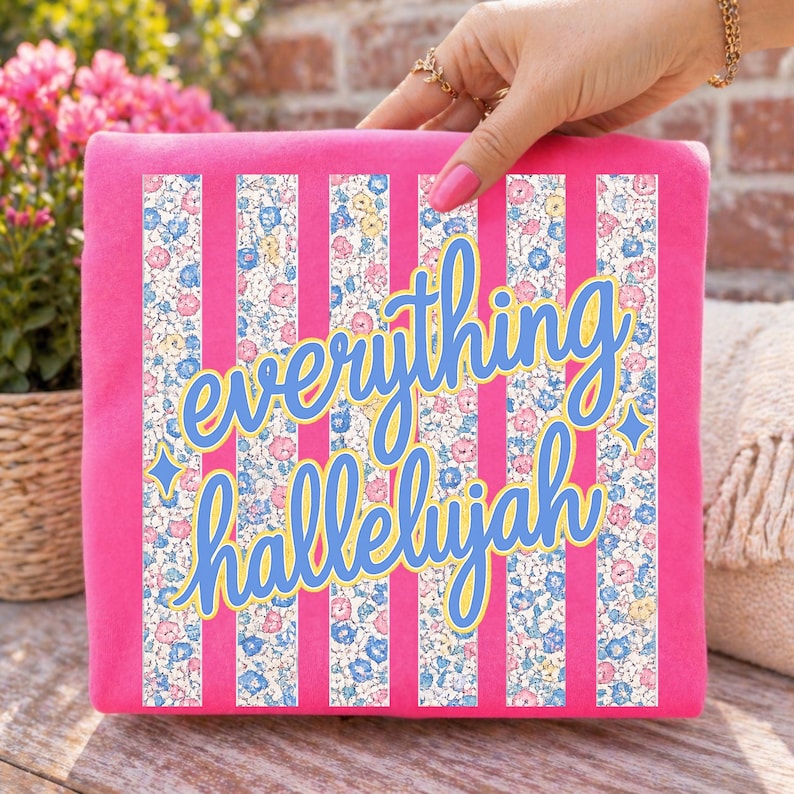 May include: A pink square decorative pillow with vertical white stripes and floral patterned stripes. The words "everything hallelujah" are written in a blue script font with a yellow outline. The pillow is held by a hand.