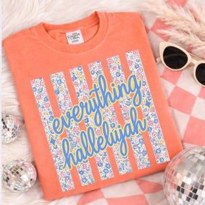 May include: An orange Comfort Colors sweatshirt with the words "everything hallelujah" in blue script. The text is set against vertical stripes filled with a floral pattern in pink, blue, and yellow.