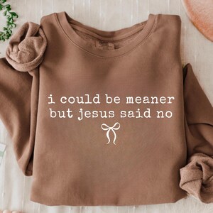 Op de afbeelding: Een bruine sweatshirt met de witte tekst "i could be meaner but jesus said no" en een witte strik. De crew neck sweatshirt is gemaakt van een zacht materiaal.