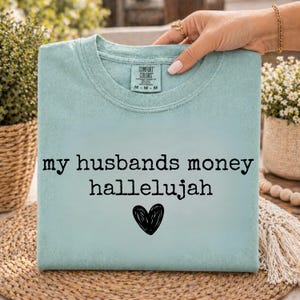 May include: A light blue crew neck t-shirt with a Comfort Colors label at the inner neckline. The front features the black text "my husbands money hallelujah" centered above a hand-drawn black heart graphic on the chest area of the garment.