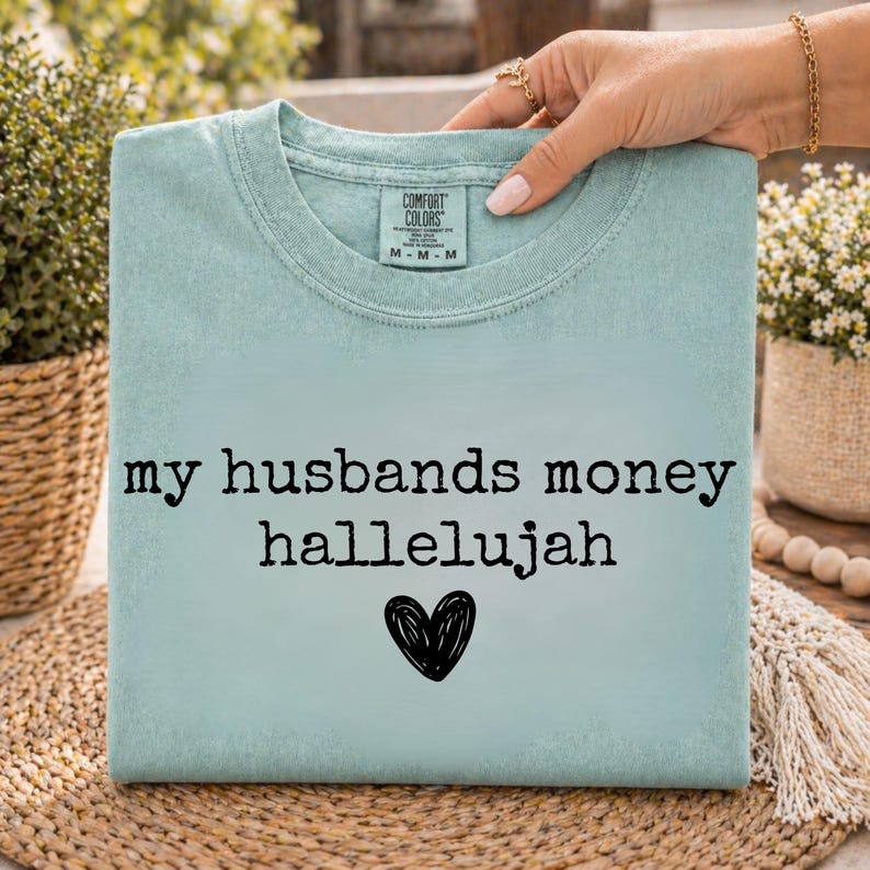 May include: A light blue crew neck t-shirt with a Comfort Colors label at the inner neckline. The front features the black text "my husbands money hallelujah" centered above a hand-drawn black heart graphic on the chest area of the garment.
