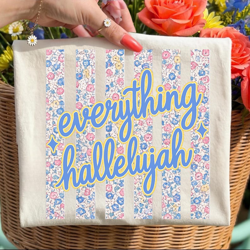 May include: Cream-colored tote bag with vertical striped floral pattern and the words "everything hallelujah" in blue script. The floral pattern includes pink, blue, and yellow flowers. The bag is in a wicker basket with flowers.