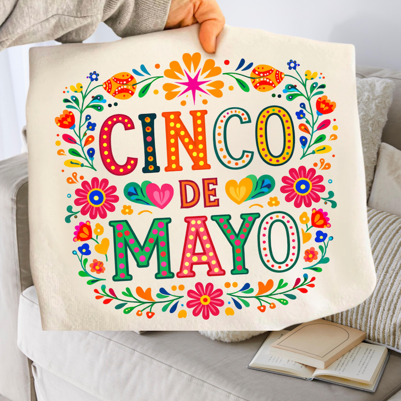 May include: A cream-coloured tote bag featuring the words "CINCO DE MAYO" in bright, decorative lettering. The text is framed by a vibrant floral wreath with red, orange, yellow, and blue accents. The bag is held up by a person.