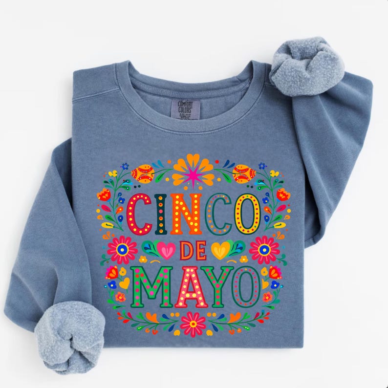 May include: A blue sweatshirt featuring the text "CINCO DE MAYO" in vibrant colours, encircled by a floral design. The sweatshirt has long sleeves with rolled-up cuffs.