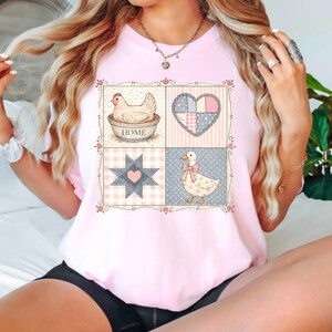 Puede incluir: Camiseta rosa claro con un dise&ntilde;o de patchwork. El dise&ntilde;o incluye una gallina en un cuenco con la palabra "HOME", un coraz&oacute;n, una estrella y un pato con un lazo. El dise&ntilde;o est&aacute; enmarcado con un borde floral.