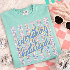 May include: A light teal t-shirt with the words "everything hallelujah" in a blue script font with a yellow outline. Vertical floral stripes in pink, blue, and yellow flowers are behind the text. Sunglasses and disco balls are in the background.