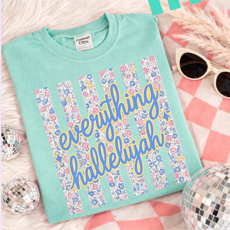 May include: A light teal t-shirt with the words "everything hallelujah" in a blue script font with a yellow outline. Vertical floral stripes in pink, blue, and yellow flowers are behind the text. Sunglasses and disco balls are in the background.