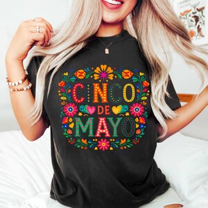May include: Black t-shirt with a vibrant Cinco de Mayo design. The design includes the words "Cinco de Mayo" in colourful lettering, framed by floral and heart motifs. The t-shirt is a relaxed, casual style.