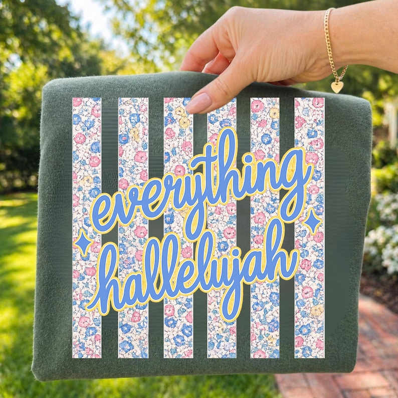 May include: A dark green rectangular item with vertical stripes of floral fabric. The text "everything hallelujah" is in blue script with a yellow outline. The floral pattern includes pink, blue, and yellow flowers.