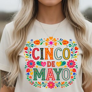 May include: A cream-coloured t-shirt with a colourful Cinco de Mayo design. The design features the words "Cinco de Mayo" in vibrant orange, green, and pink, surrounded by a floral wreath with flowers and leaves.