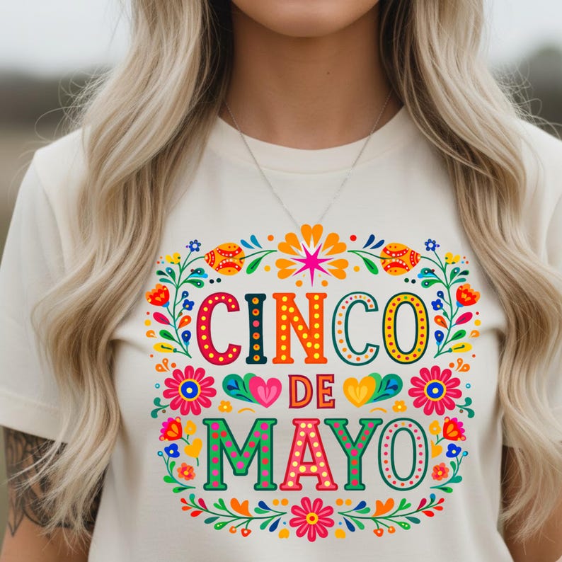 May include: A cream-coloured t-shirt with a colourful Cinco de Mayo design. The design features the words "Cinco de Mayo" in vibrant orange, green, and pink, surrounded by a floral wreath with flowers and leaves.
