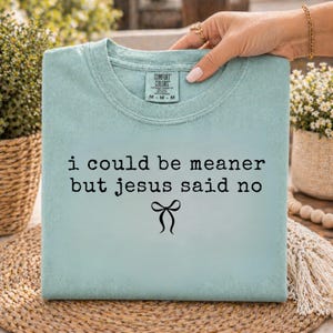 Op de afbeelding: Lichtblauw T-shirt met de zwarte tekst "i could be meaner but jesus said no" en een strik-afbeelding. Het T-shirt wordt vastgehouden door een hand met gouden sieraden.